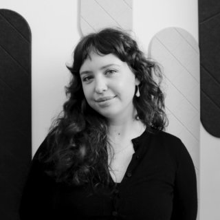 Portrait of Ava Tyrrell Walsh - Receptionist at MOY wearing a dark top, standing indoors against a textured wall, looking at the camera with a slight smile.